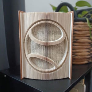 Könnte beinhalten: Eine Buchfaltkunst-Skulptur mit einem Tennisball-Design, das durch präzises Falten der einzelnen Seiten eines Hardcover-Buches entsteht. Die cremefarbenen Papierseiten sind so geformt, dass sie die Kreisform und die geschwungenen Linien bilden.