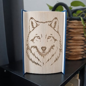 Peut inclure: Une sculpture en livre représentant une tête de loup créée en pliant les pages d'un livre. La sculpture possède une couverture bleu foncé et présente un portrait détaillé et symétrique d'une tête de loup formé par les bords des pages.