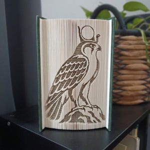 May include: A folded book art sculpture featuring the intricate silhouette of an Egyptian falcon deity, likely Horus, with a sun disk crown. The pages are precisely creased to form the bird's profile, feathers, and base, creating a three-dimensional effect.