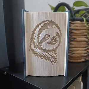 Könnte beinhalten: Eine Buchfaltkunst-Skulptur, die ein detailliertes Faultiergesicht zeigt, welches durch präzises Falten der einzelnen Seiten eines Hardcover-Buches entsteht. Die cremefarbenen Seitenkanten bilden das Bild, während die dunkelblauen Buchdeckel sichtbar sind.
