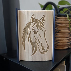 Pode incluir: Uma escultura de arte em livro dobrado apresentando uma silhueta detalhada de uma cabeça de cavalo criada dobrando com precisão as páginas individuais de um livro de capa dura. O livro tem uma capa azul escura e páginas creme com texto.