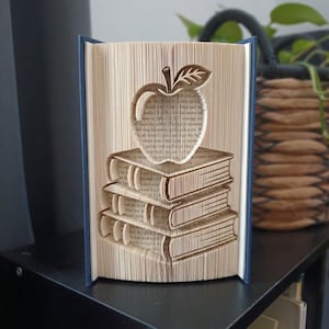 May include: A book sculpture featuring folded pages that form the shape of an apple resting on a stack of three books. The book has a dark blue cover, and the interior pages are cream-colored with visible printed text throughout the folded design.