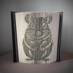 May include: A unique book art piece featuring a gray chinchilla design. The chinchilla is created by precisely folding the pages of a book. The book is displayed upright, with the cut design visible, and has a dark gray spine and covers.