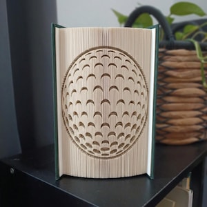 May include: A folded book art sculpture featuring a three-dimensional golf ball design created by precisely folding the individual pages of a hardback book with a dark green spine. The pages are arranged to form a spherical, dimpled pattern.