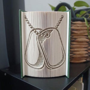 Peut inclure: Une sculpture en livre avec des pages pliées formant la silhouette de deux plaques d'identité militaires suspendues à une chaîne à billes. Le livre possède une couverture vert foncé et les pages intérieures sont blanches avec du texte imprimé.