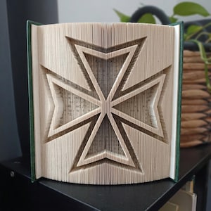 May include: A folded book art sculpture featuring a Maltese cross design created by precisely folding the individual pages of a hardback book. The book has a dark green cover and the pages are arranged to form a sharp, symmetrical cross shape.