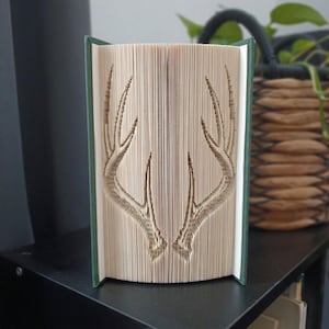 May include: A folded book art sculpture featuring a symmetrical design of two deer antlers created by precisely folding the individual pages of a hardback book with a dark green cover, standing upright to display the intricate paper craft pattern.