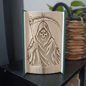 May include: A folded book art sculpture featuring a detailed silhouette of a grim reaper holding a scythe. The pages are precisely creased to form the figure, with the book covers visible in dark green on the left and right edges of the sculpture.