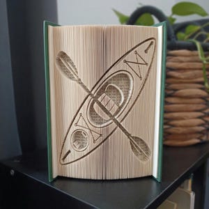 May include: A book art sculpture featuring a folded page design that creates the silhouette of a kayak and a paddle. The book has a dark green cover and the pages are cream-coloured, showing the intricate folding technique used to form the shape.