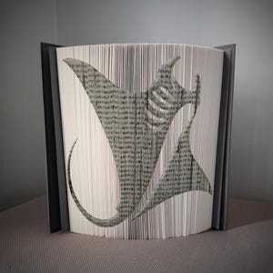 May include: A unique book art sculpture featuring a manta ray design. The pages are folded and cut to create the shape of the marine animal. The book has a gray cover and the pages are a light beige color.