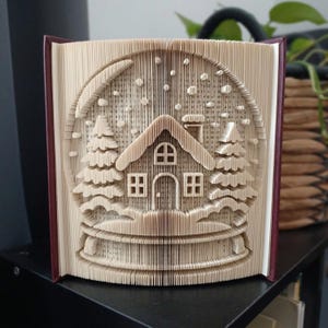 Può includere: Una scultura in libro piegato che raffigura una scena invernale con una casa centrale, due pini e una luna crescente. Le pagine sono piegate con precisione per formare un effetto palla di neve tridimensionale con accenti di neve che cade.
