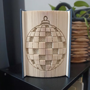 May include: A folded book art sculpture featuring a three-dimensional Christmas ornament design created by precisely folding the individual pages of a hardback book. The beige paper pages are manipulated to form a spherical bauble shape.