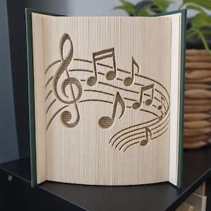 May include: A folded book art sculpture featuring a treble clef and musical notes carved into the pages. The book has a dark green cover and the pages are arranged to create a three-dimensional musical staff design with visible paper edges.