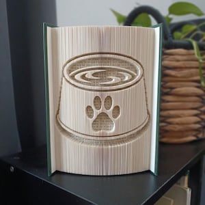May include: A folded book art sculpture featuring a pet food bowl design with a paw print cutout on the front. The pages are precisely folded to create the three-dimensional shape of a bowl filled with food, set against a dark green book cover.