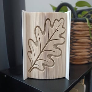 Op de afbeelding: Een boeksculptuur met gevouwen pagina's die de duidelijke vorm van een eikenblad vormen. De randen van het papier zijn nauwkeurig gevouwen om een driedimensionaal silhouet van het blad te creëren, met zichtbare tekst op de pagina's.