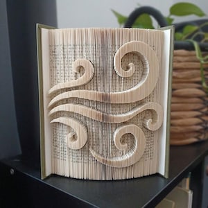 Puede incluir: Una escultura de libro creada doblando las páginas de un libro de tapa dura para formar un patrón simétrico de cuatro formas onduladas y giratorias. Los bordes del papel color crema están plegados con precisión para dar textura.