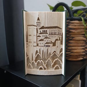 May include: A book sculpture featuring intricate folded pages that form the silhouette of a castle with a tall central tower, multiple levels of buildings, and arched windows, all created through precise paper folding techniques.