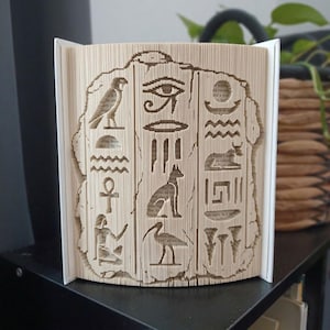 May include: A folded book art sculpture featuring intricate cutouts of ancient Egyptian symbols including the Eye of Horus, an ankh, a jackal, and various hieroglyphic figures. The pages are precisely folded to create a three-dimensional relief.