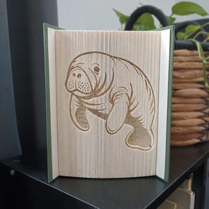Op de afbeelding: Een boeksculptuur met een lamantijnontwerp, gemaakt door de pagina's van een hardcover boek te vouwen. De lamantijn is in profiel afgebeeld met zichtbare gezichtsdetails en vinnen, gevormd door de nauwkeurige rangschikking van de papierranden.
