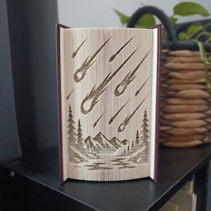 Peut inclure: Une sculpture en livre plié présentant un motif découpé en papier représentant une pluie de météores tombant sur une chaîne de montagnes et des pins. Les pages sont pliées avec précision pour créer un paysage en trois dimensions.