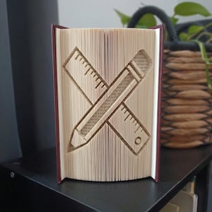 Puede incluir: Una escultura de libro plegado que presenta un diseño en forma de cruz de una regla y un lápiz. Las páginas están dobladas y marcadas con precisión para crear la silueta de estos dos artículos de papelería sobre el papel color crema.