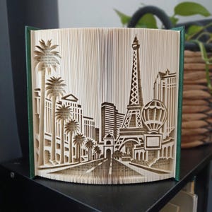 Op de afbeelding: Een gevouwen boekkunstsculptuur met een gedetailleerd uit papier gesneden stadsgezicht van Las Vegas. Het ontwerp bevat een replica van de Eiffeltoren, palmbomen en een heteluchtballon, allemaal gemaakt door de pagina's van het boek te vouwen.
