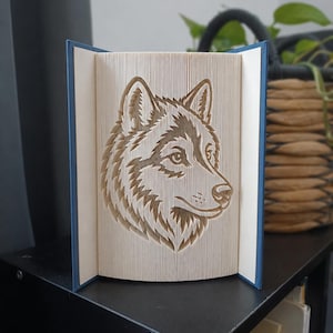May include: A book sculpture featuring a folded page art design of a wolf head in profile. The book has a blue cover and the pages are precisely folded to create the detailed image of the wolf, showing its ears, snout, and fur texture clearly.