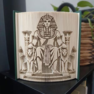 May include: A book sculpture featuring a folded page design of an Egyptian pharaoh sitting on a throne flanked by two lion statues. The pages are precisely creased to create a three-dimensional image of the figure wearing a traditional headdress.