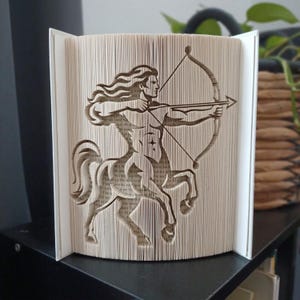 May include: A folded book art sculpture featuring the silhouette of a centaur archer. The pages are precisely creased to form the image of a figure with a human torso and horse body, holding a bow and arrow. The paper edges are cream-coloured.