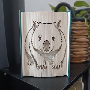 Peut inclure: Une sculpture de livre représentant un wombat, réalisée en pliant les pages d'un livre à couverture rigide. Le wombat est centré sur les pages ouvertes, montrant son visage, ses oreilles et ses pattes avant. Le livre a des couvertures bleu sarcelle.