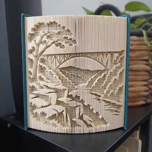 Könnte beinhalten: Eine Buchskulptur aus gefalteten Seiten, die eine Landschaftsszene bilden. Das Design zeigt eine große Bogenbrücke über einem Fluss, mit einem markanten Baum auf der linken Seite und felsigem Gelände entlang der Ufer. Das Buch hat einen blaugrünen Einband.