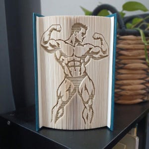 Peut inclure: Une sculpture en livre plié représentant la silhouette détaillée d'un culturiste musclé posant avec les bras fléchis. Les pages sont pliées avec précision pour créer l'image, avec une couverture bleu foncé, posée sur une surface.
