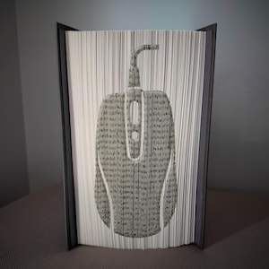 Pode incluir: Uma peça de arte de livro única com um design de rato de computador. A silhueta do rato é criada dobrando cuidadosamente as páginas de um livro. O livro tem uma capa cinza escuro e as páginas são cinza claro.