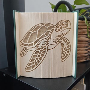 May include: A folded book art sculpture featuring a detailed sea turtle design created by precisely folding the pages of a hardcover book. The turtle is depicted in profile with visible shell patterns and flippers, showcasing the paper edges.