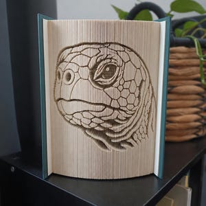 May include: A book sculpture featuring a folded page art design of a sea turtle head in profile. The pages are precisely creased to form the detailed texture of the turtle's scales, eye, and snout, with teal-coloured hardcovers on both sides.