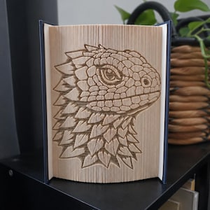 Peut inclure: Une sculpture en livre avec un design de pages pliées formant le profil détaillé d'une tête de lézard ou de dragon aux écailles texturées. Le livre est ouvert avec des couvertures bleu foncé et se tient debout, révélant le pliage complexe.