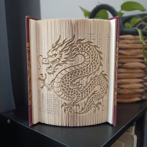 Peut inclure: Une sculpture de livre représentant un dragon créée en pliant les pages d'un livre à couverture rigide. Le dragon est représenté de profil avec des écailles complexes, un long corps serpentin et des traits détaillés formés par les bords.