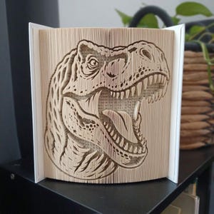 May include: A book sculpture featuring a folded page art design of a Tyrannosaurus Rex head. The pages are precisely folded to create the three-dimensional silhouette of the dinosaur, showing its open mouth, sharp teeth, and textured skin.