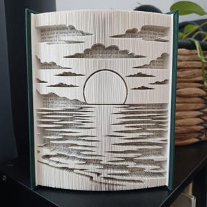 May include: A folded book art sculpture featuring a sunset scene with a circular sun, layered clouds, and a reflective water surface created by precisely folding the individual pages of a hardcover book with dark green covers.