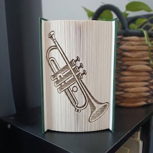 May include: A folded book art sculpture featuring a trumpet design created by precisely folding the individual pages of a hardback book. The trumpet shape is clearly defined by the edges of the paper, showcasing the instrument's valves and bell.