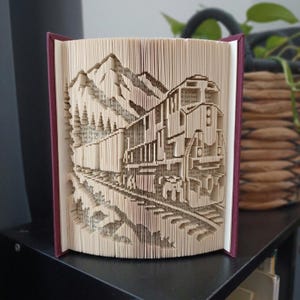 May include: A folded book art sculpture featuring a detailed train locomotive traveling along tracks through a mountainous landscape. The pages are precisely creased to form the three-dimensional image, with dark maroon covers visible on the sides.