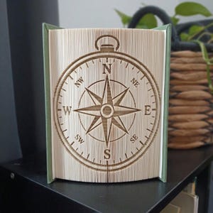 Puede incluir: Una escultura de libro plegado que presenta un diseño detallado de rosa de los vientos creado doblando las páginas con precisión. La brújula muestra las direcciones cardinales e intercardinales N, NE, E, SE, S, SW, W y NW con una cubierta verde.