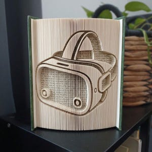 Könnte beinhalten: Eine Buchskulptur mit gefalteten Seiten, die die Form eines Virtual-Reality-Headsets bilden. Die Seiten sind cremefarben mit sichtbarem schwarzem Textdruck, und das Buch hat einen dunkelgrünen Einband an den linken und rechten Rändern.