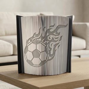 Peut inclure: Sculpture de livre décoratif avec un ballon de football et des flammes découpés dans les pages. Le livre ouvert, avec une tranche noire, est posé sur une table en bois. Le motif est visible sur les bords des pages.