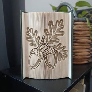 Könnte beinhalten: Eine Buchfaltkunst-Skulptur mit einem detaillierten Ausschnittdesign von zwei Eicheln mit Eichenblättern. Die Seiten sind präzise gefaltet und geknickt, um das dreidimensionale Bild der Früchte und des Laubs an den weißen Papierkanten zu erzeugen.