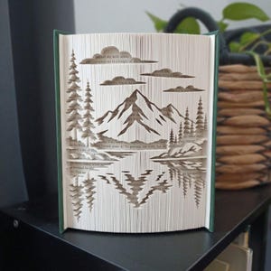 Puede incluir: Una escultura de libro con páginas dobladas con precisión que crean un paisaje tridimensional. El diseño muestra un pico de montaña central, árboles de hoja perenne circundantes y nubes, con un efecto de reflejo en el agua.