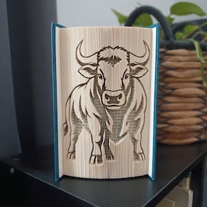 Op de afbeelding: Een gevouwen boekkunstsculptuur met een gedetailleerd stierontwerp, gemaakt door de pagina's van een boek nauwkeurig te vouwen. De stier is van voren afgebeeld met prominente hoorns en gespierde kenmerken, tegen een donkerblauwe kaft.