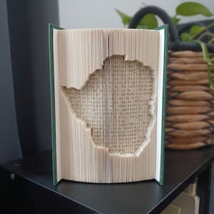 Peut inclure: Une sculpture de livre avec des pages pliées formant le contour distinct du pays du Zimbabwe. Le livre possède une couverture vert foncé et est placé à la verticale, mettant en valeur la technique complexe de pliage de papier utilisée.