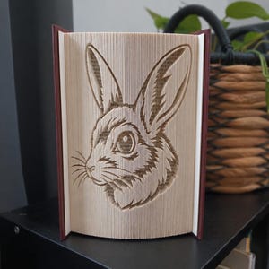May include: A folded book art sculpture featuring a detailed rabbit head design created by precisely folding the individual pages of a hardcover book. The rabbit is shown in profile with long ears, whiskers, and a textured fur appearance.
