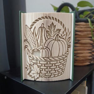 Puede incluir: Una escultura de libro plegado con temática de cosecha. Las páginas están dobladas intrincadamente para crear un diseño tridimensional de una cesta tejida llena de una calabaza, un calabacín, una mazorca de maíz y hojas de arce, sobre una cubierta verde.